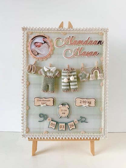 Personalized birth-details keepsake board with a circular baby photo, a tiny crocheted onesie and pants clipped to a mini clothesline, and wooden birth-date, weight, and time tags