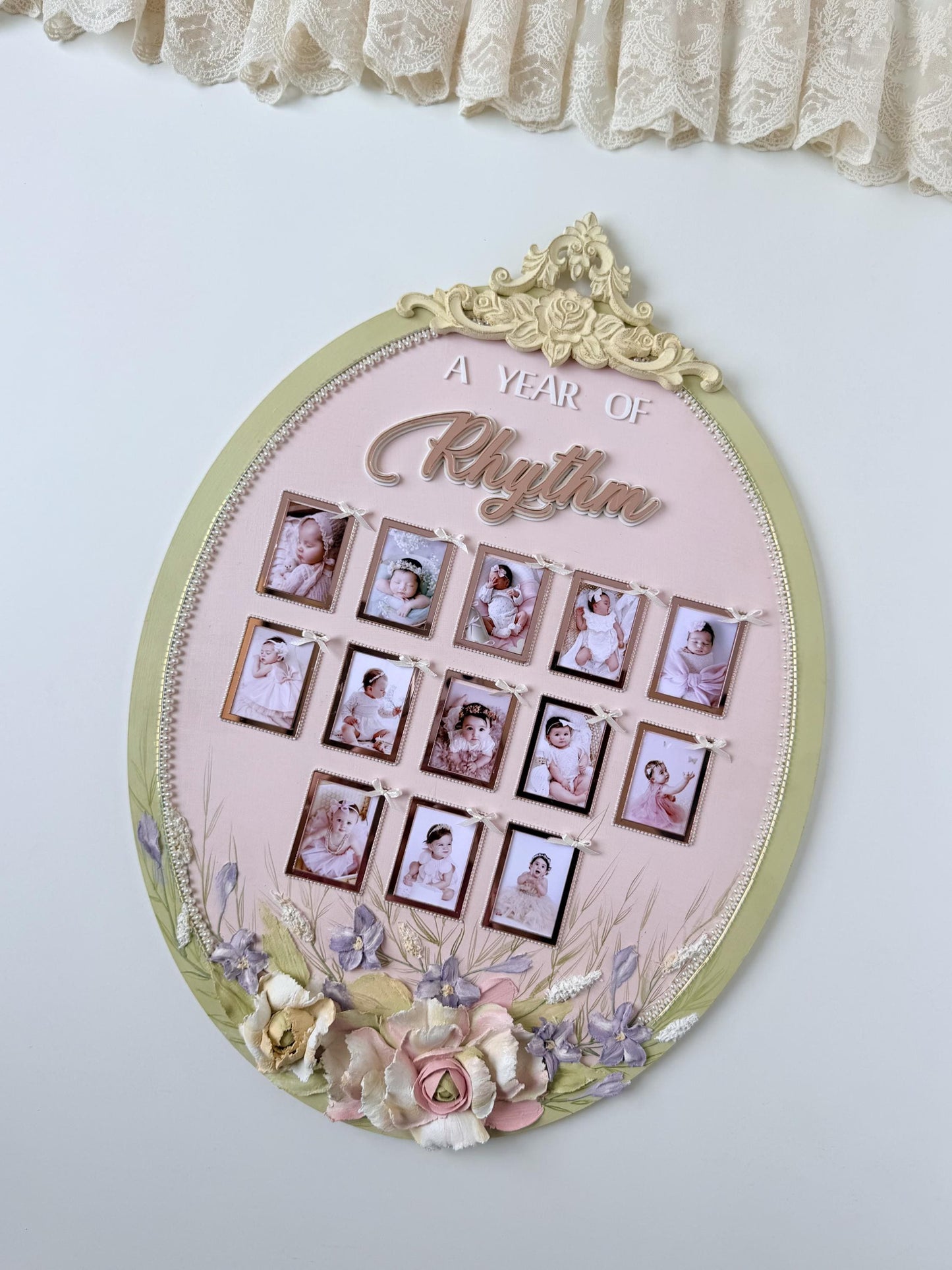 Oval nursery keepsake frame with a floral crest and thirteen ribbon-tied photo frames tracking a baby's first year, newborn through one year