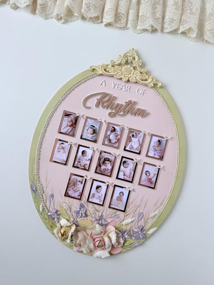 Oval nursery keepsake frame with a floral crest and thirteen ribbon-tied photo frames tracking a baby's first year, newborn through one year
