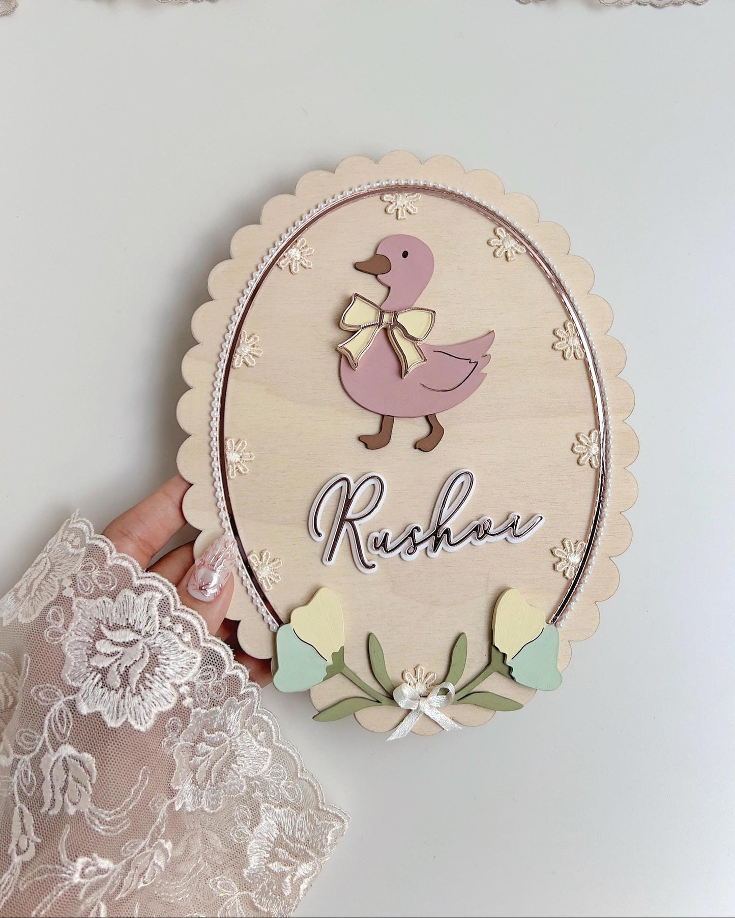 Personalized wooden oval nursery sign with a pastel-pink duck design, the baby's name in script, and lace and pearl trim, displayed on a small easel